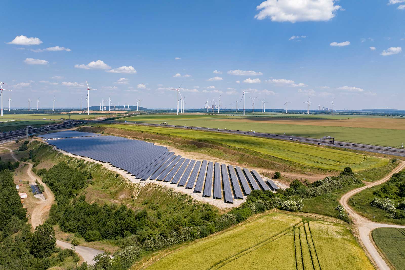 Luftaufnahme von Solarmodulen auf einem Hügel, mit Windkraftanlagen in der Ferne und Feldern unter einem hellblauen Himmel.
