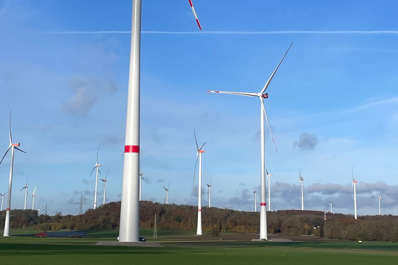Eine Landschaft mit mehreren Windkraftanlagen vor einem strahlend blauen Himmel und grünen Feldern.