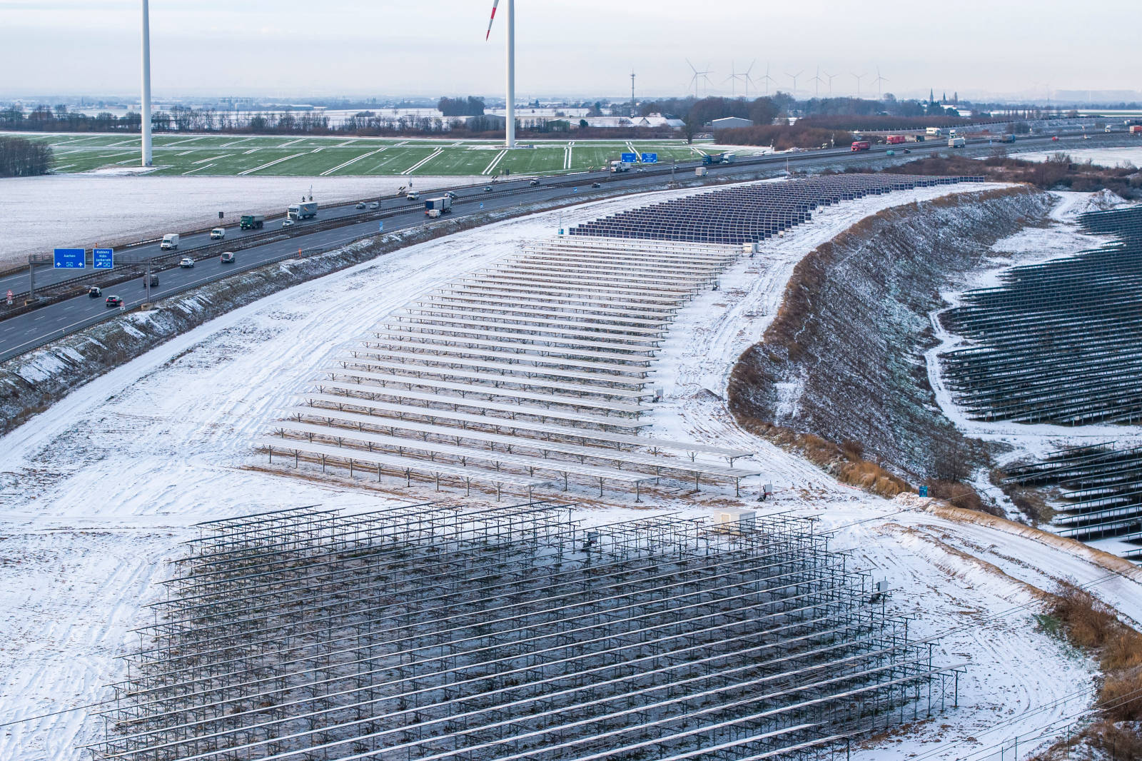 Eine verschneite Landschaft mit Solarpanels und Windkraftanlagen neben einer belebten Straße.