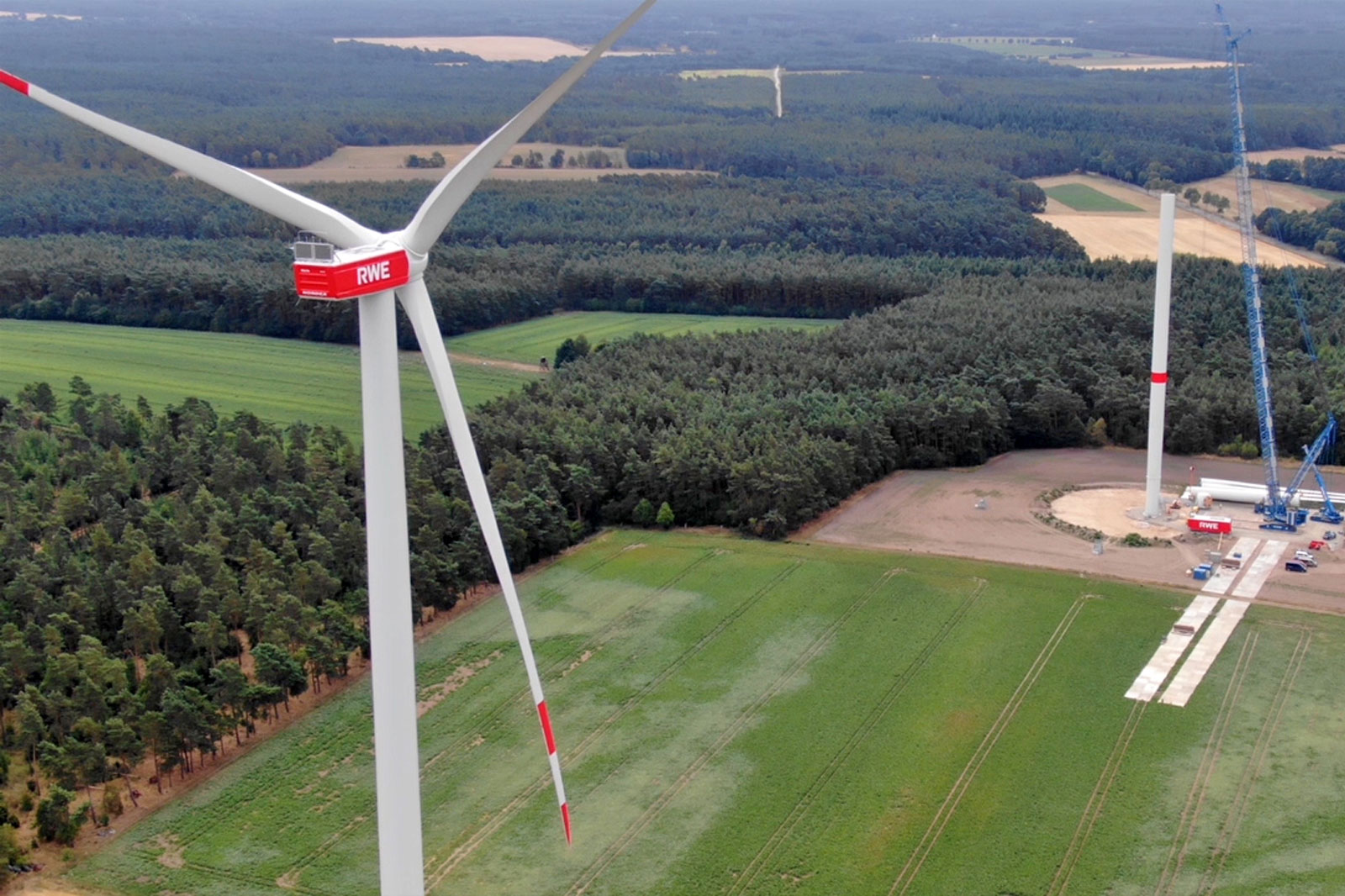 Eine Luftaufnahme eines Windparks mit großen Turbinen inmitten grüner Felder und eines Waldes.