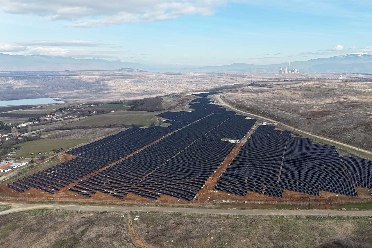 A large solar farm with numerous solar panels stretches across the landscape under a blue sky.