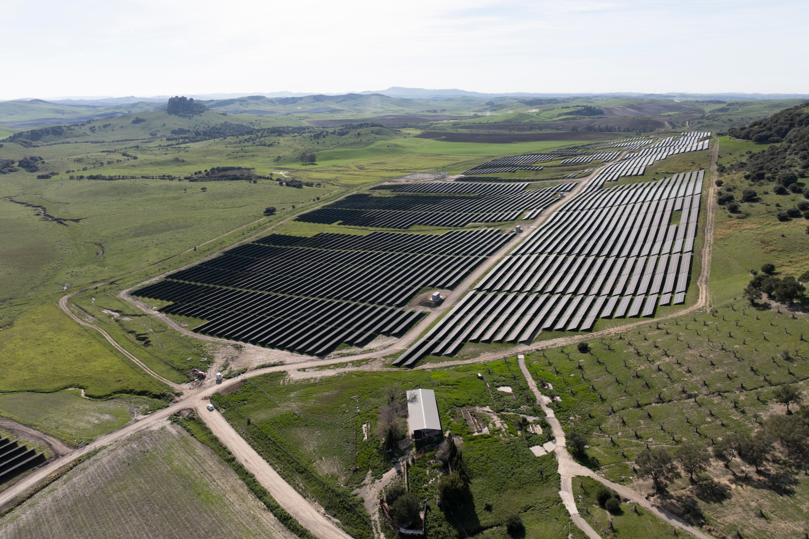 Aerial view of a large solar farm with rows of solar panels, surrounded by green fields and hills in the background.