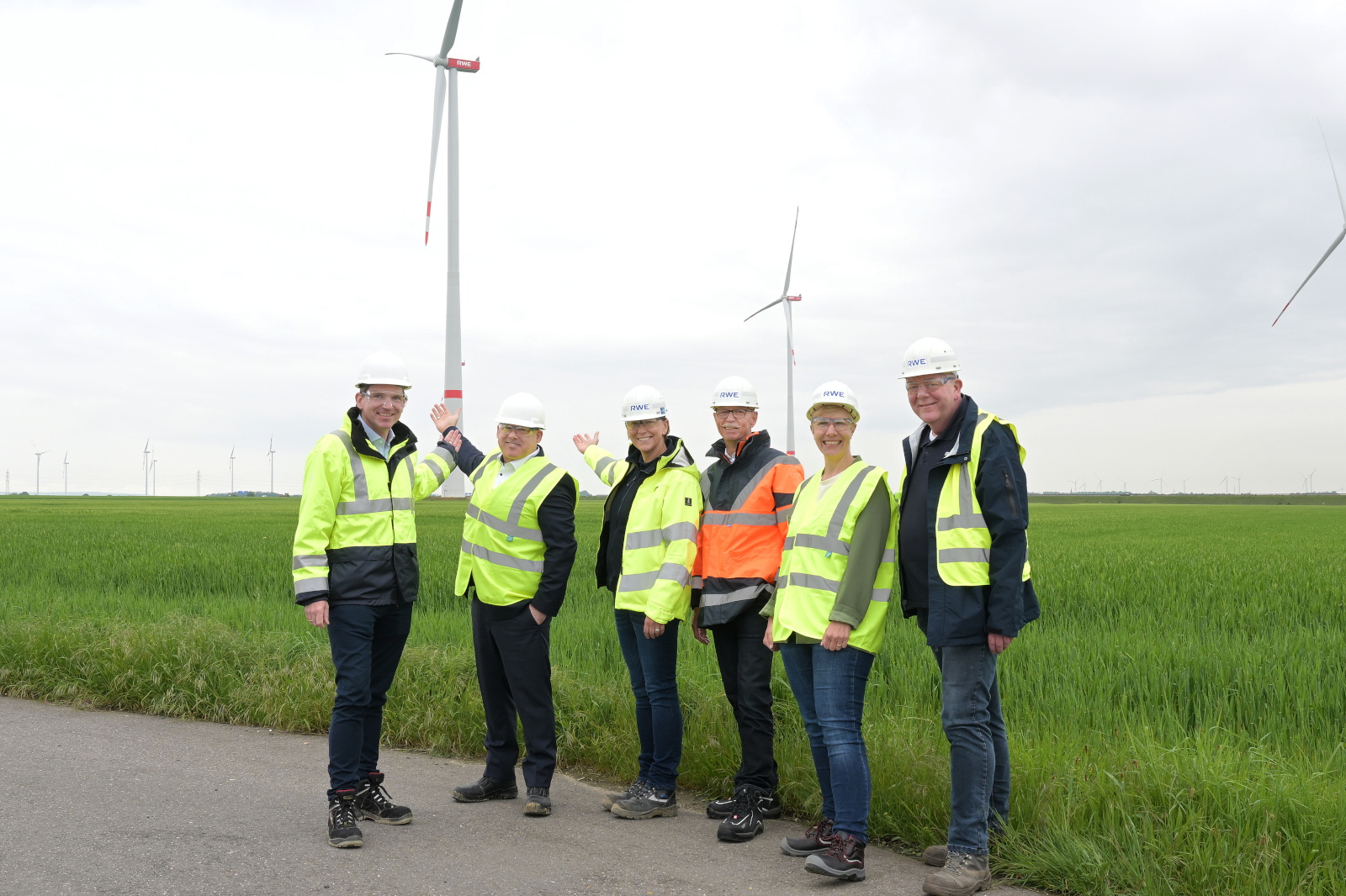 A group of six individuals in safety clothing stands in front of a wind turbine in a green field.