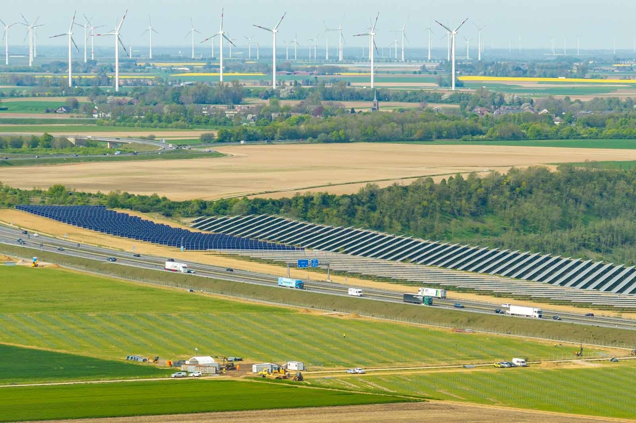 Eine malerische Sicht mit Solarpanelen neben einer Autobahn, Windkraftanlagen im Hintergrund und Feldern in der Ferne.