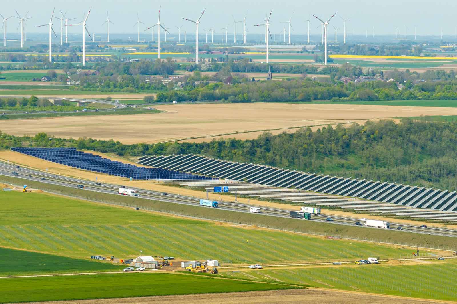 Eine malerische Sicht mit Solarpanelen neben einer Autobahn, Windkraftanlagen im Hintergrund und Feldern in der Ferne.