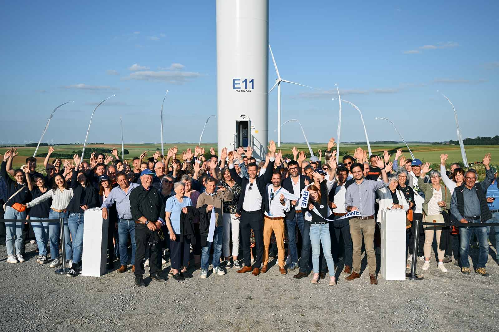 A large group of people standing in front of a wind turbine, smiling and waving with a clear sky in the background.