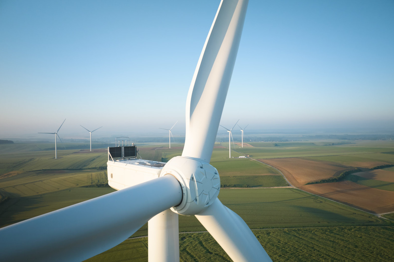 Close-up of a wind turbine blade with multiple turbines in the background, set against a clear blue sky and green fields.