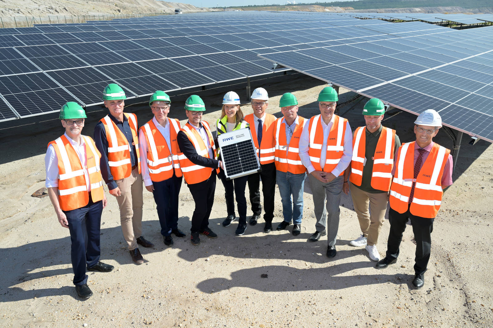 A group of people wearing safety vests and helmets stands near solar panels, holding a solar panel.