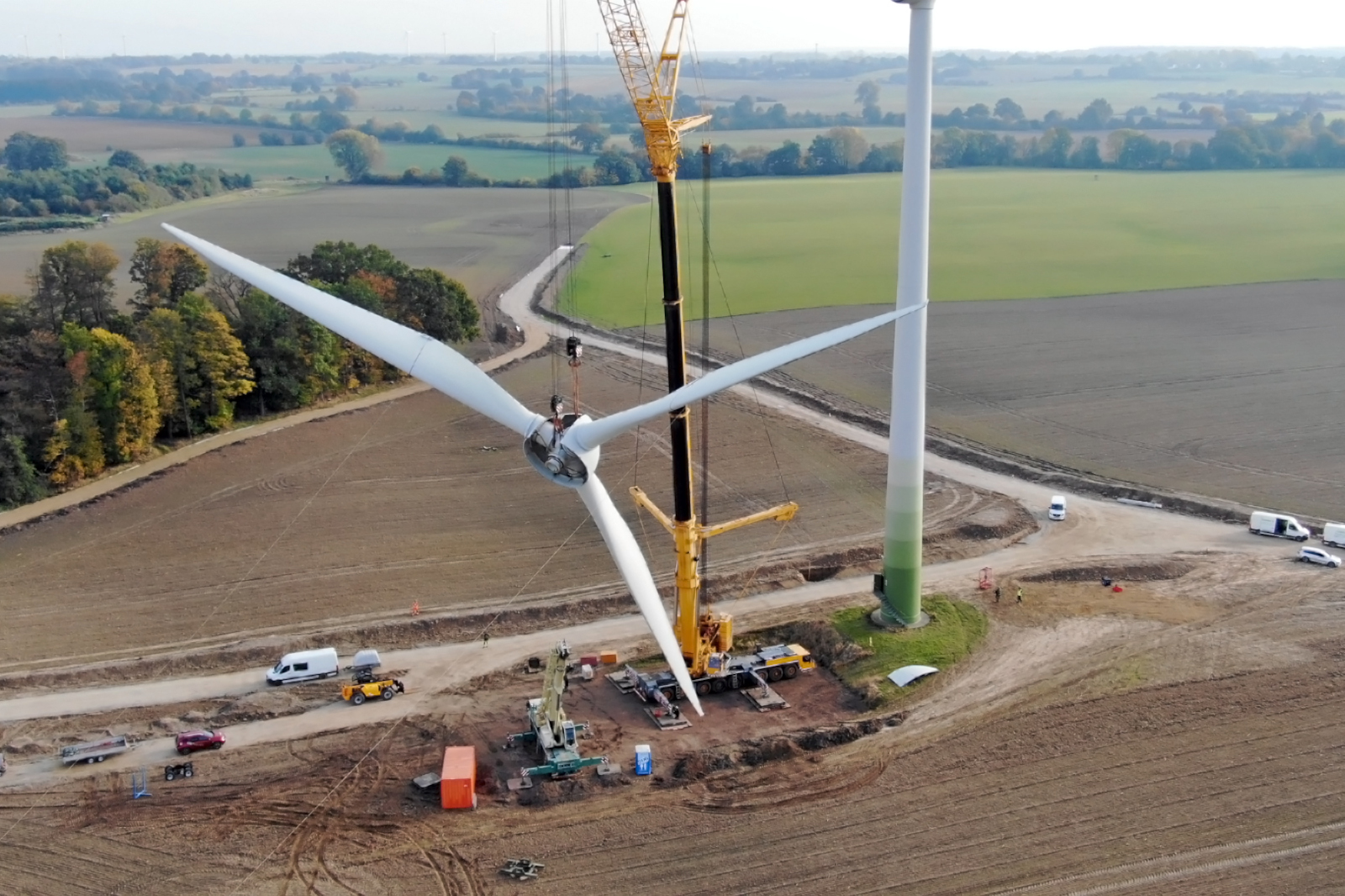 A crane lifts the blade tip of a wind turbine over a field for installation. Construction vehicles are visible.
