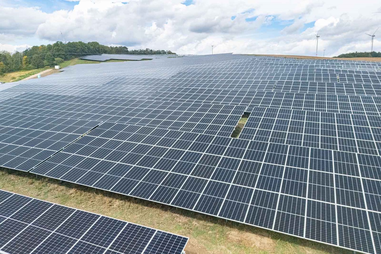 Eine große Fläche mit Solarpanels, die in einem hügeligen Gelände installiert sind, umgeben von Bäumen und einem blauen Himmel.