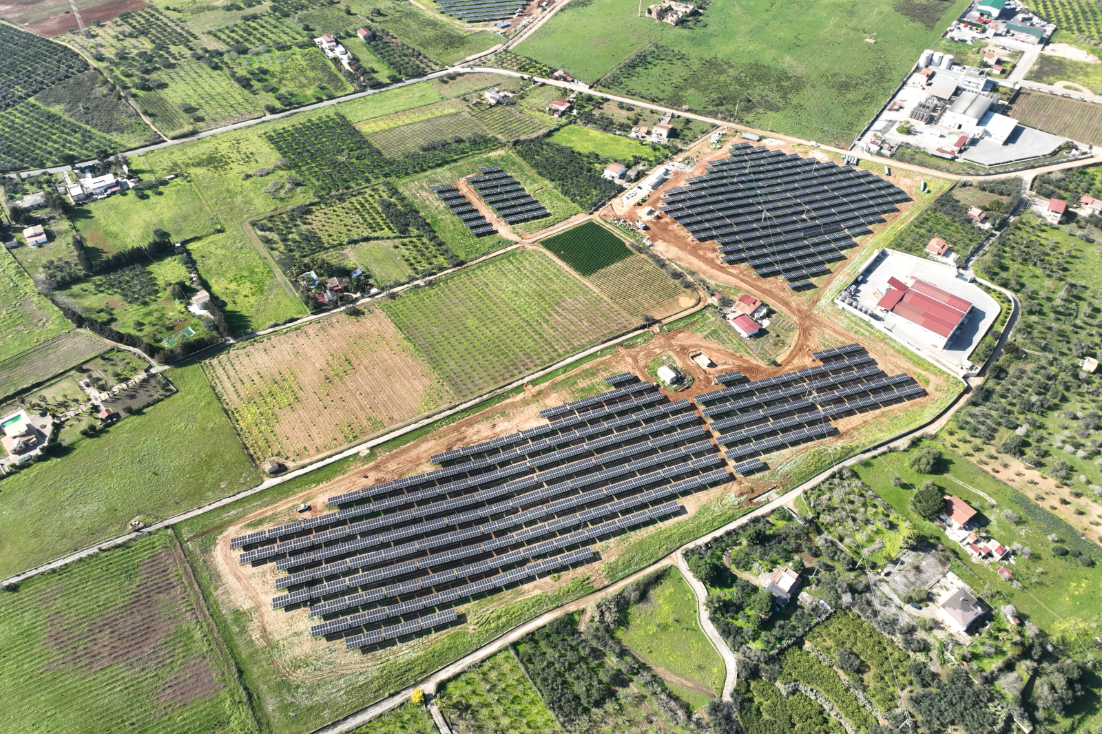 An aerial view of a solar farm with rows of solar panels surrounded by green fields and scattered buildings.