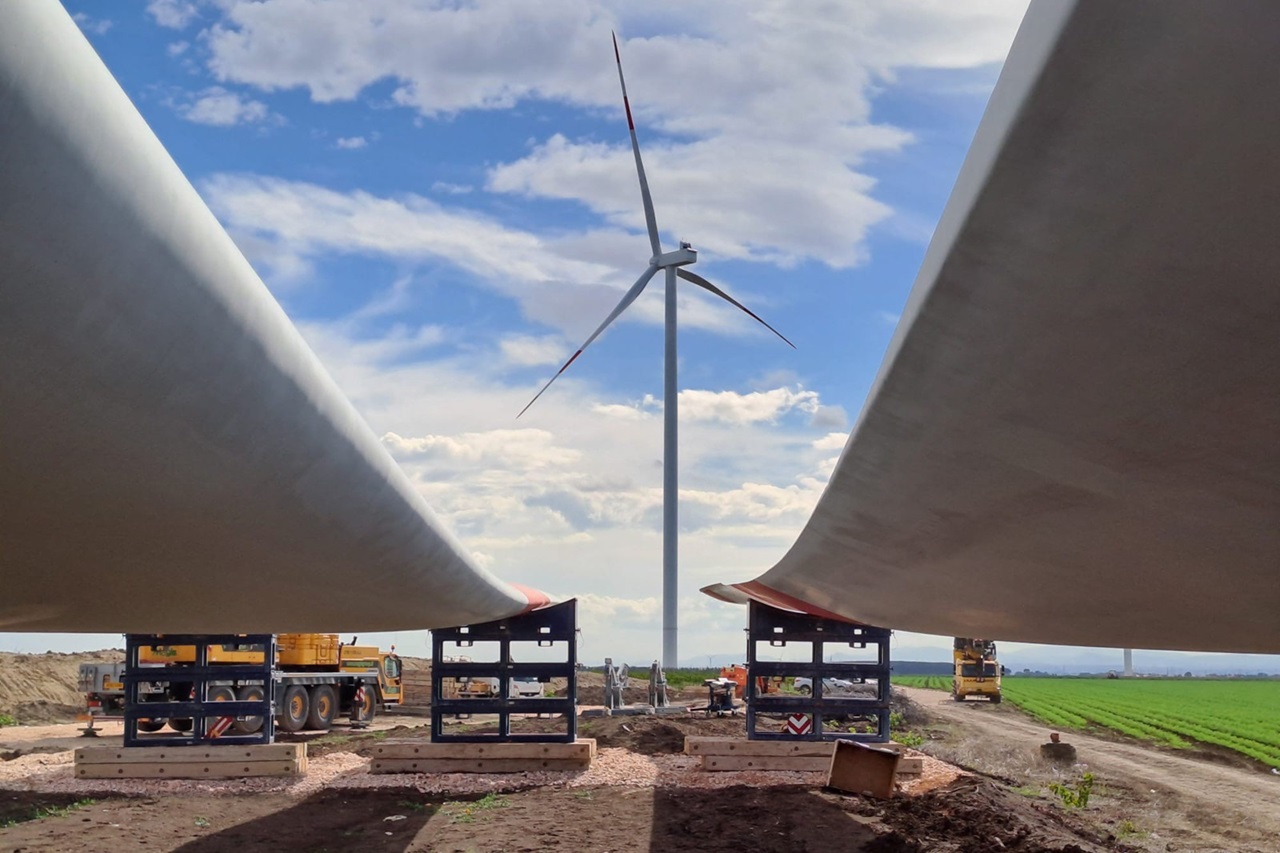 Two wind turbine blades are positioned on cranes with a wind turbine in the background against a blue sky with clouds.