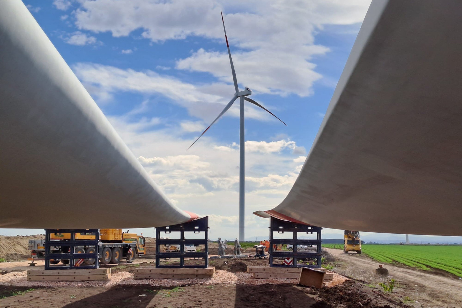 Two wind turbine blades are positioned on cranes with a wind turbine in the background against a blue sky with clouds.
