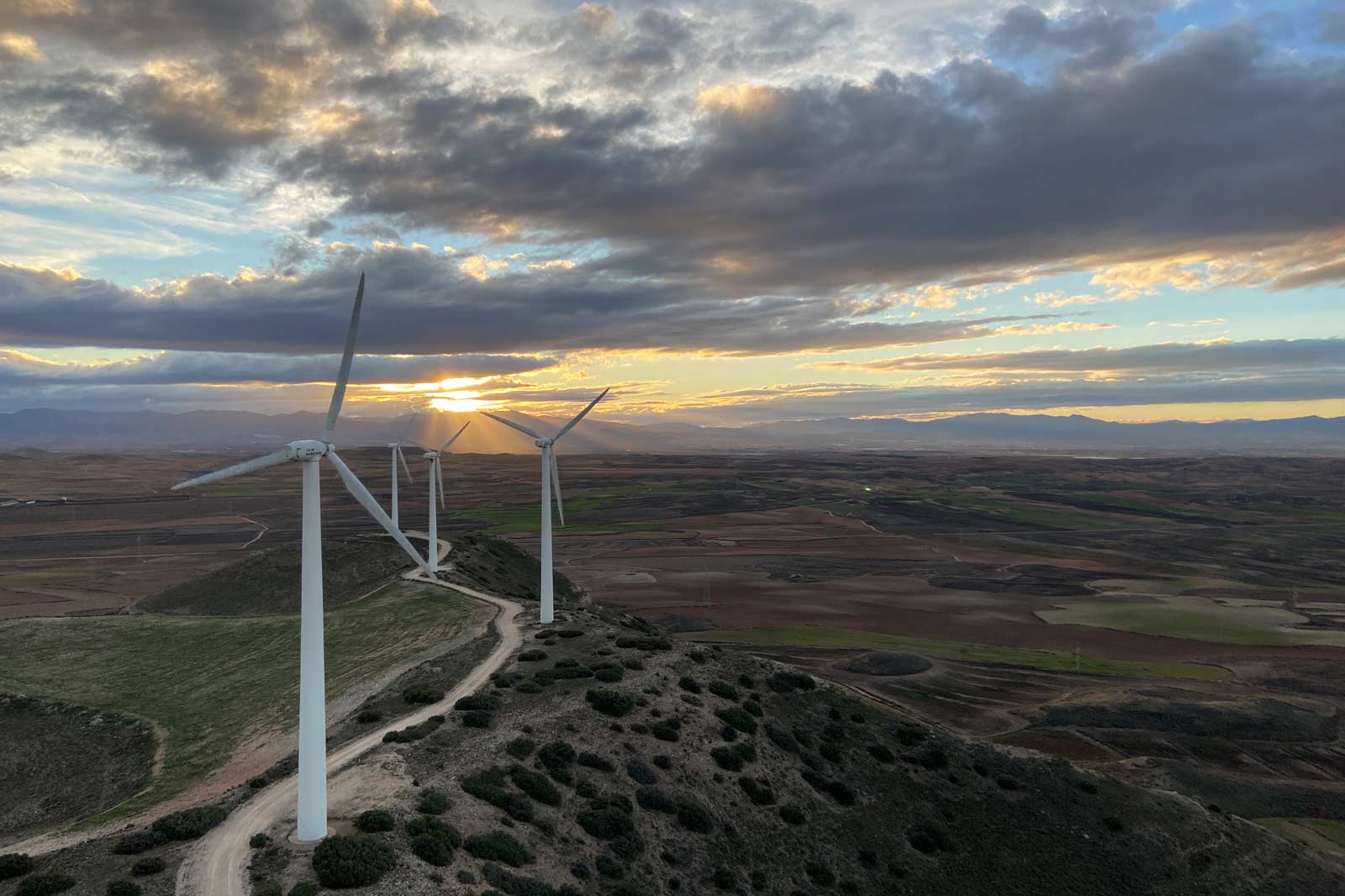 Windkraftanlagen stehen auf einem Hügel mit einer Landschaft unter einem dramatischen Himmel bei Sonnenuntergang.