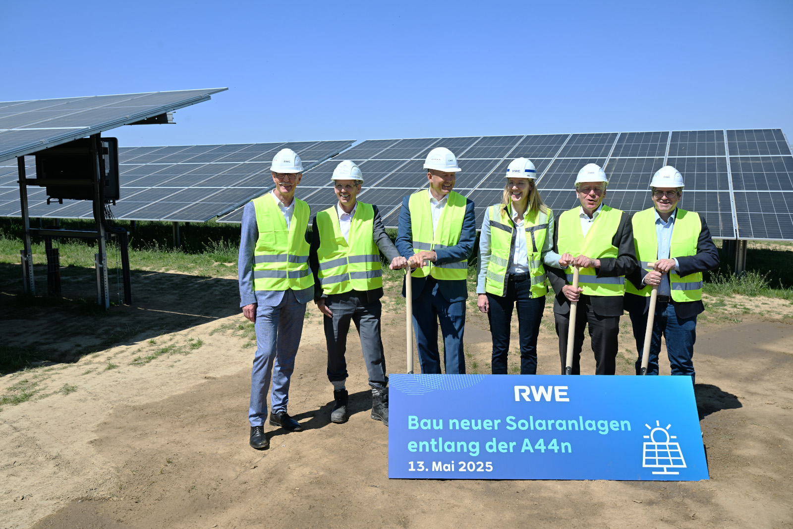 Sechs Personen in Sicherheitswesten und Helmen stehen vor Solarpanelen. Sie halten Schaufeln und eine Tafel mit Informationen.