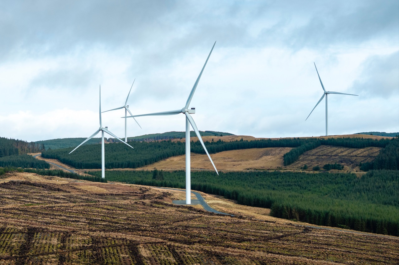 A landscape with several wind turbines on a hillside, surrounded by green forests and cloudy skies.