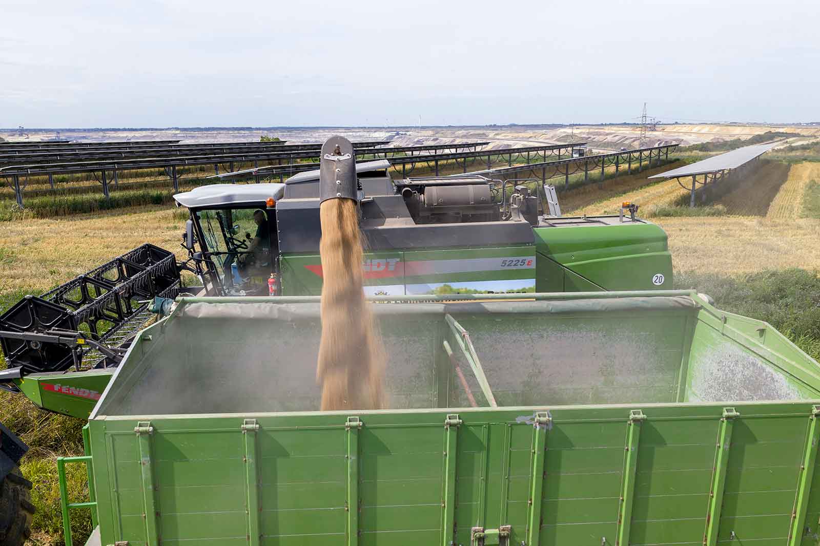 A combine harvester unloads grain into a green trailer. Solar panels and fields are visible in the background.