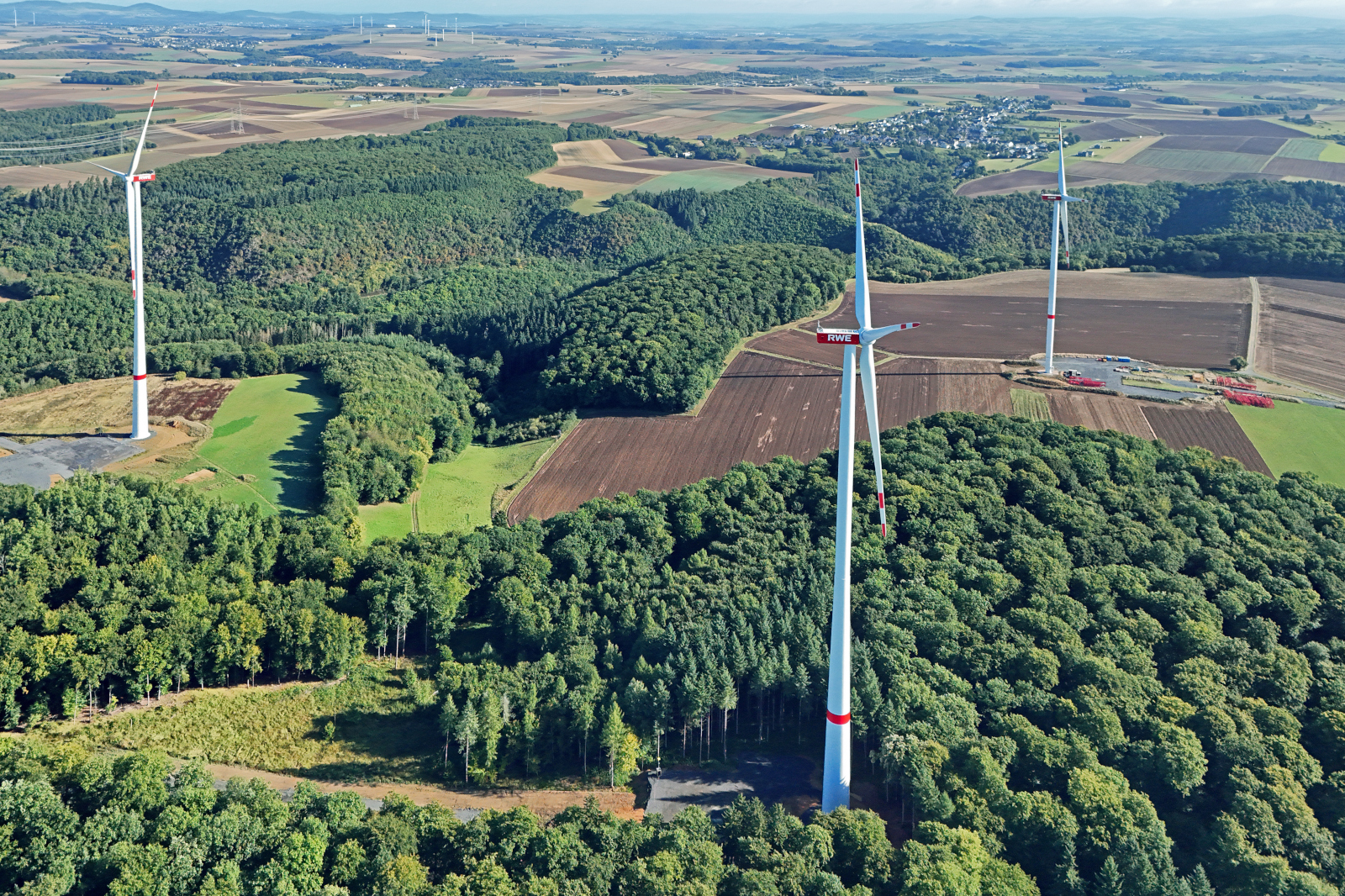 Luftaufnahme von Windkraftanlagen zwischen grünen Wäldern und landwirtschaftlichen Feldern unter einem klaren Himmel.