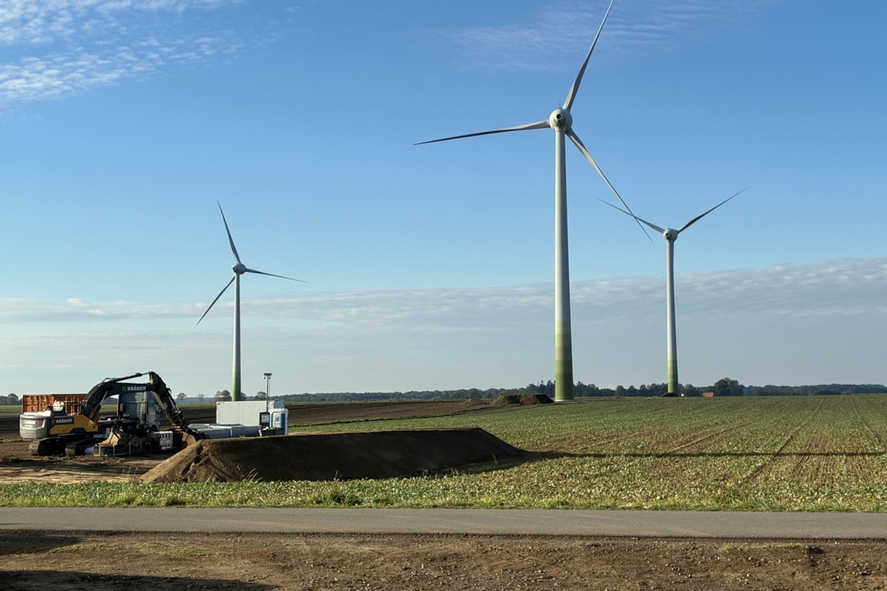 Drei Windkraftanlagen stehen in einem Feld, während ein Bagger Erde bewegt und eine Baufläche vorbereitet.