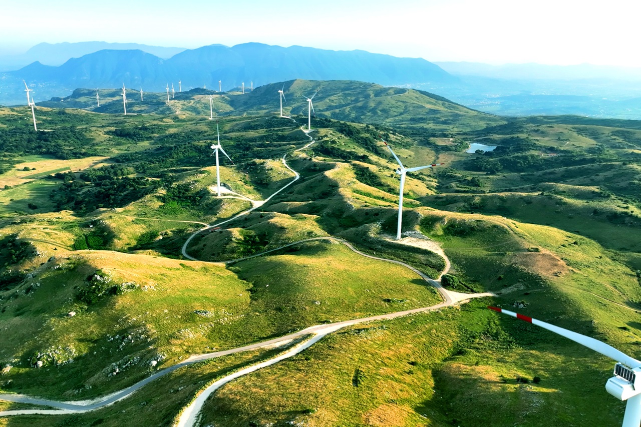Luftaufnahme einer hügeligen Landschaft mit Windkraftanlagen und kurvigen Straßen, umgeben von grünen Wiesen.