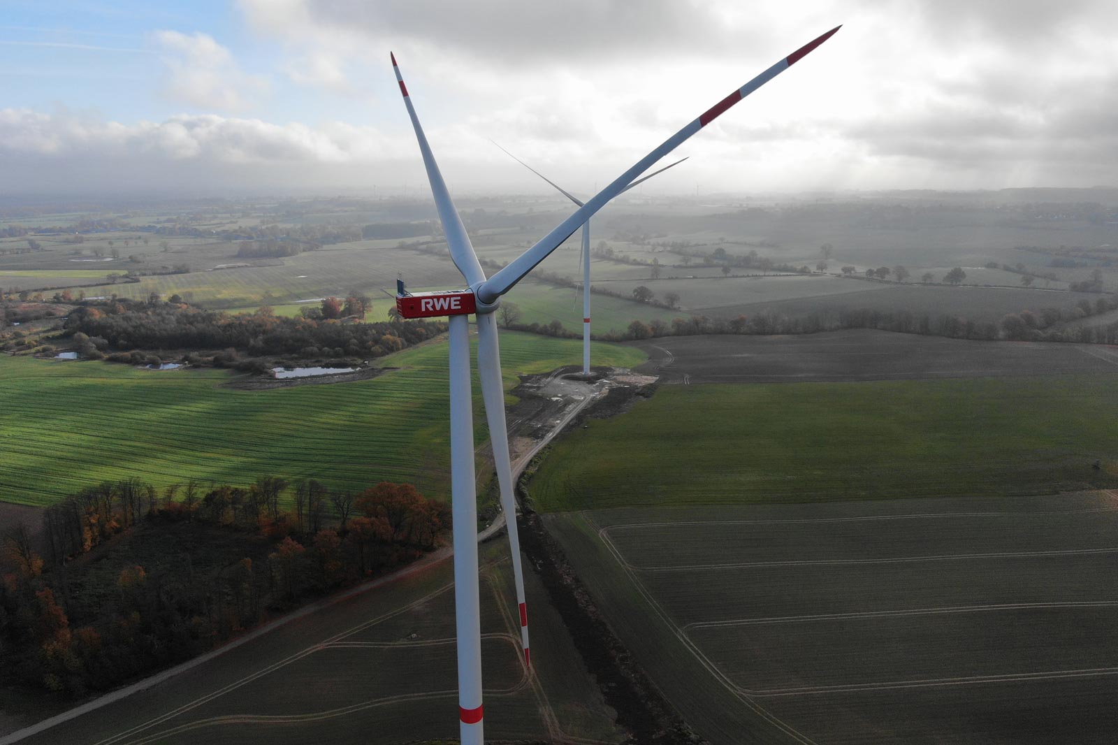 Ein Windrad von RWE steht über grünen Feldern, umgeben von Bäumen und Wolken, in einer ländlichen Umgebung.