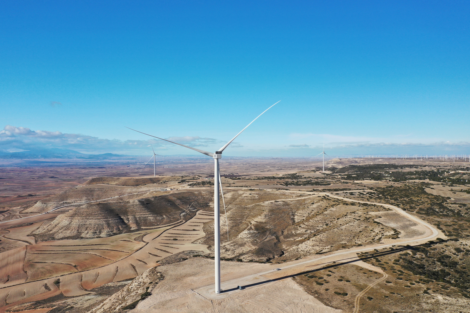 A wind turbine stands on hilly terrain with gentle earth waves and a clear blue sky in the background.