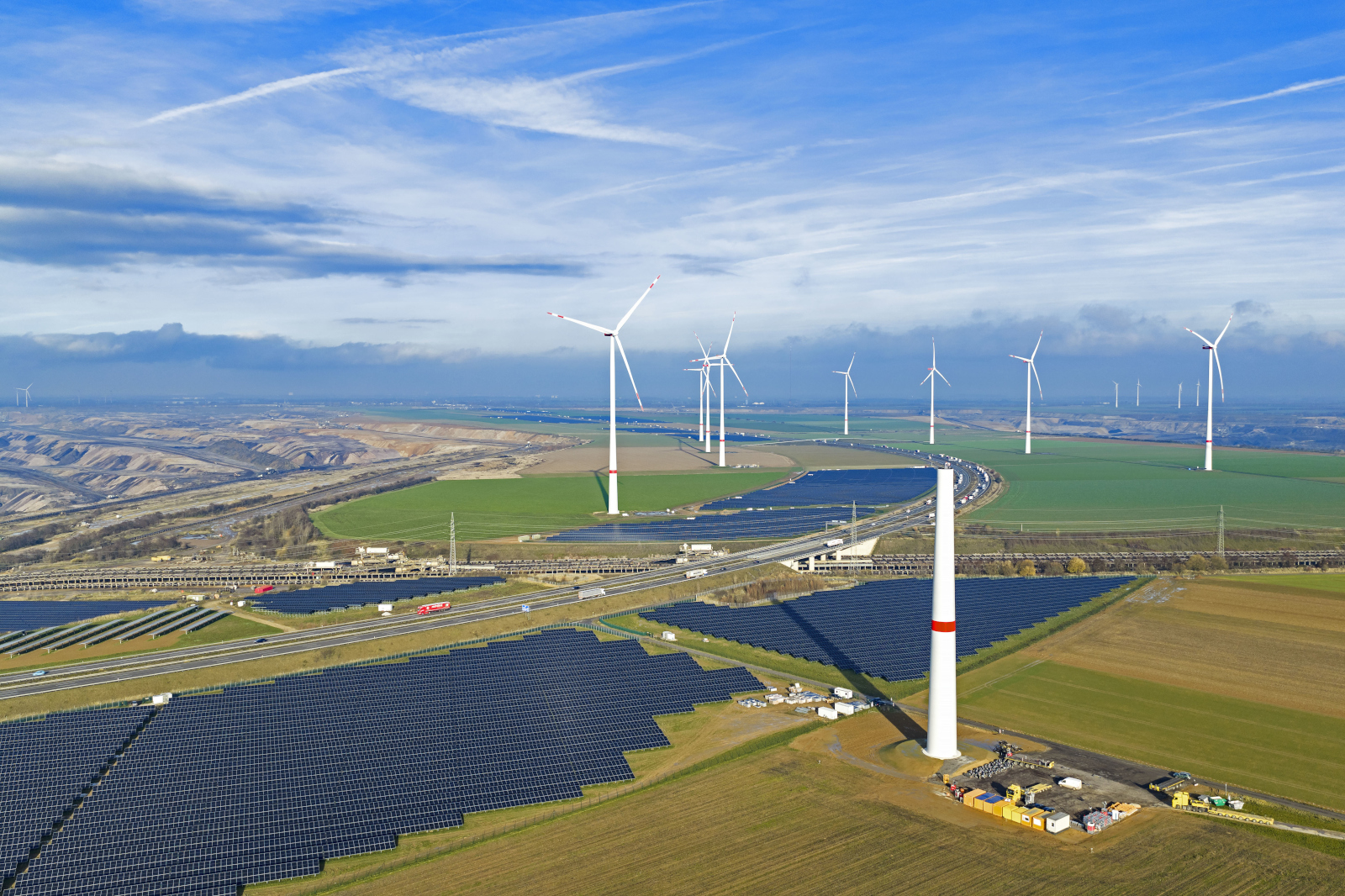Eine Luftaufnahme zeigt Windturbinen und Solarpanelle, die in einer weitläufigen, grünen Landschaft angeordnet sind.