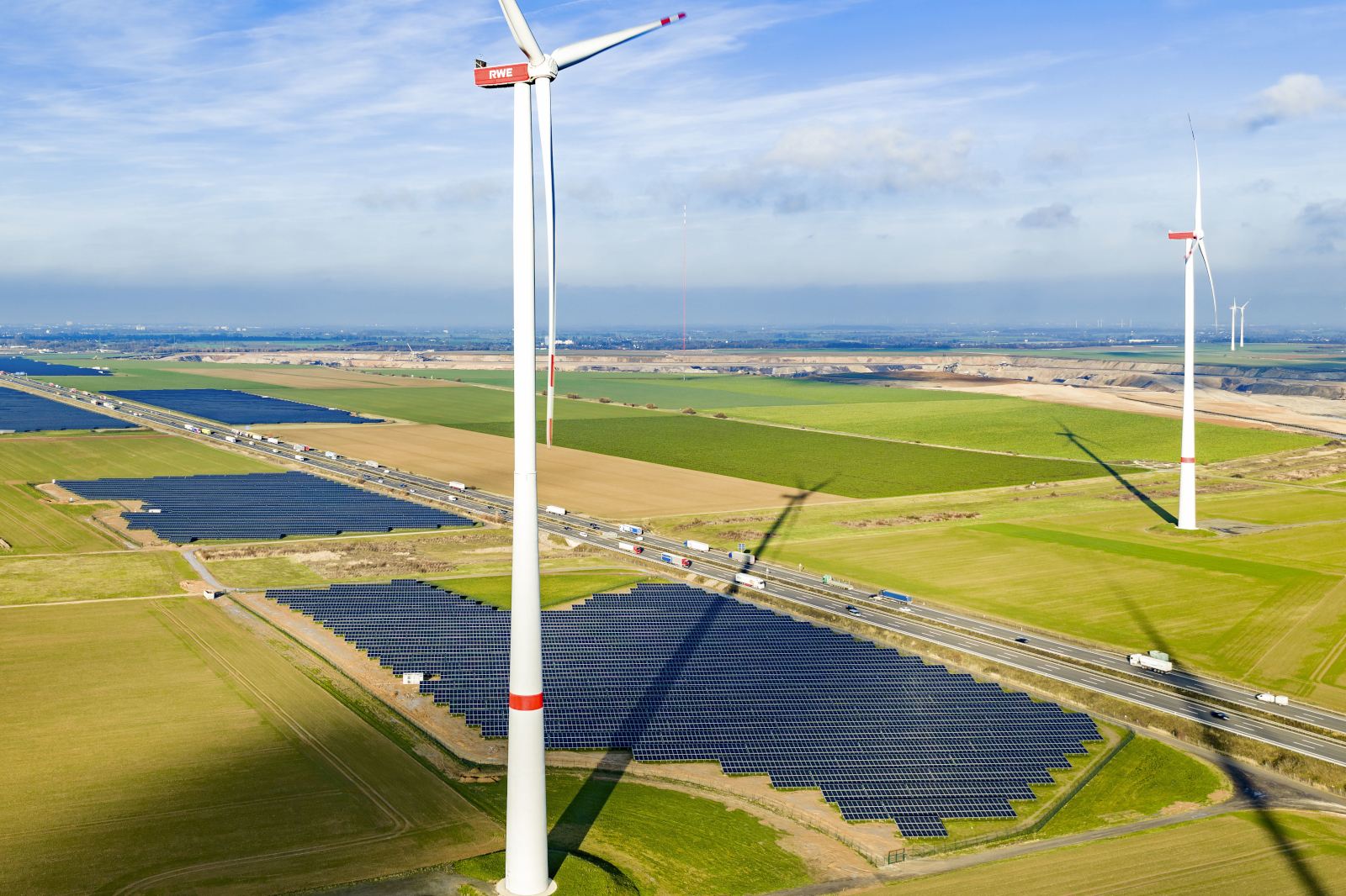 Eine Windturbine steht neben einem großen Solarpark, mit der Autobahn und grünen Feldern im Hintergrund.