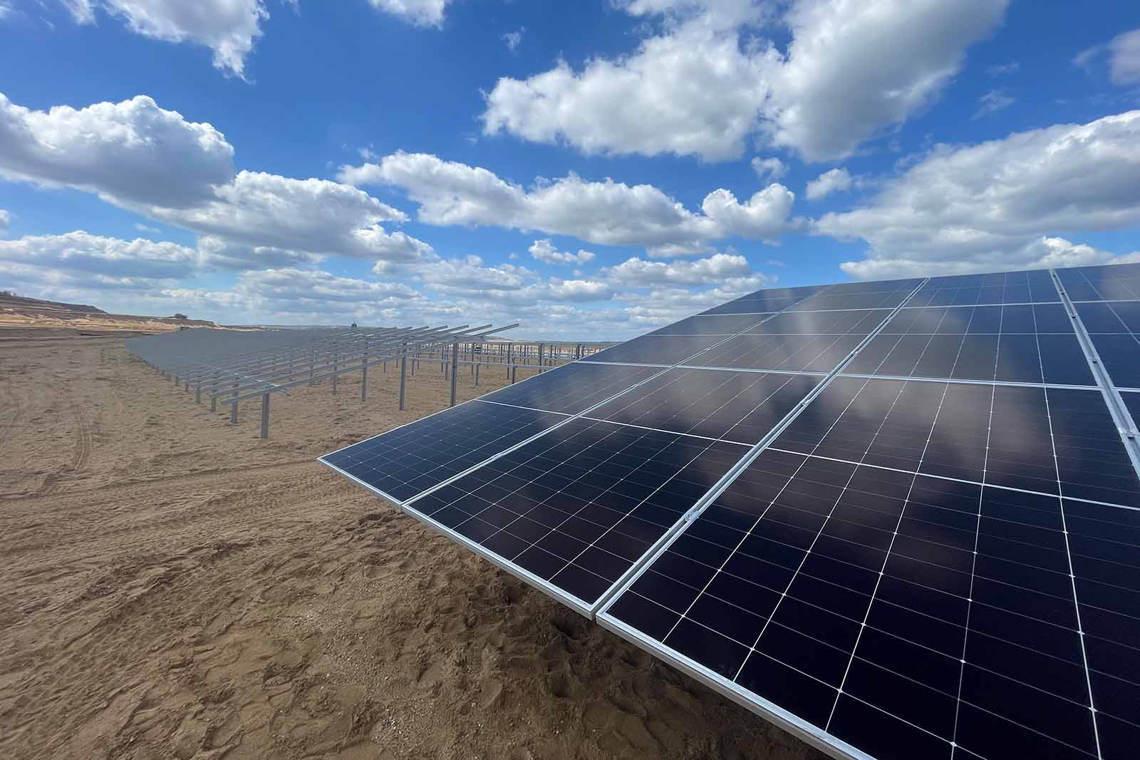 Glossy dark blue solar panels with silver frames fill the foreground on sandy ground, with rows stretching toward a bright blue sky and white clouds.