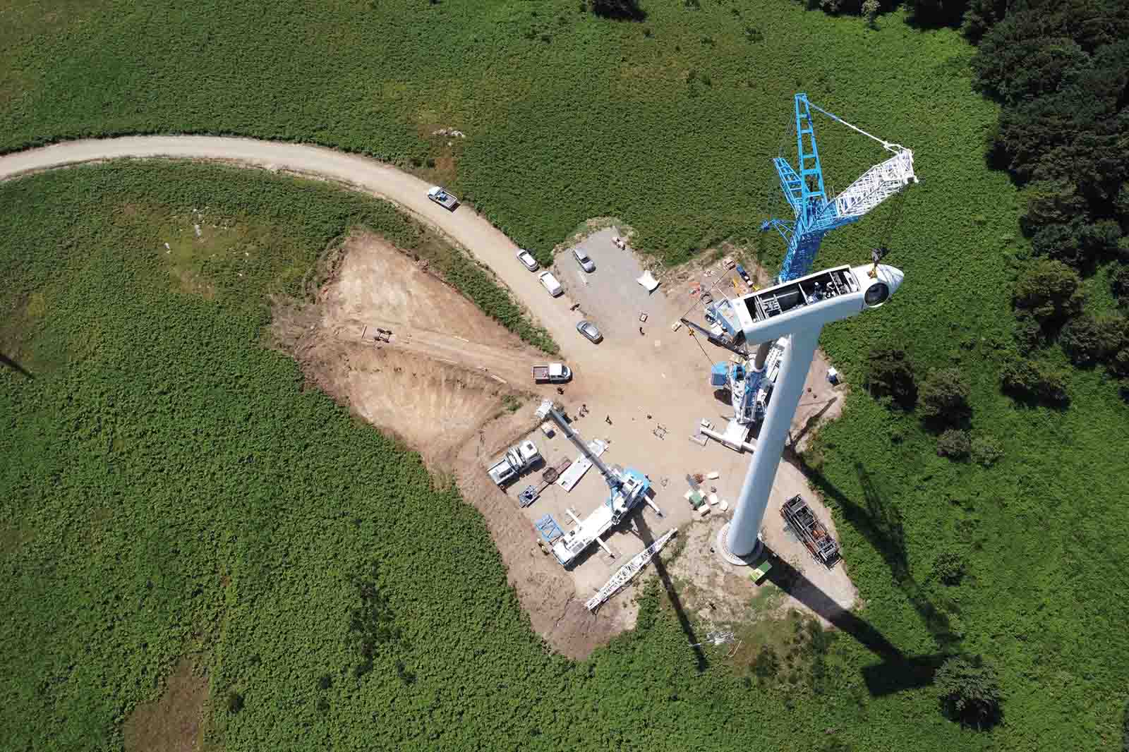 Weißer zylindrischer Turm mit Gondel steht neben einem blauen Kran auf einer Erdfläche, umgeben von grünen Feldern und mehreren weißen Fahrzeugen.