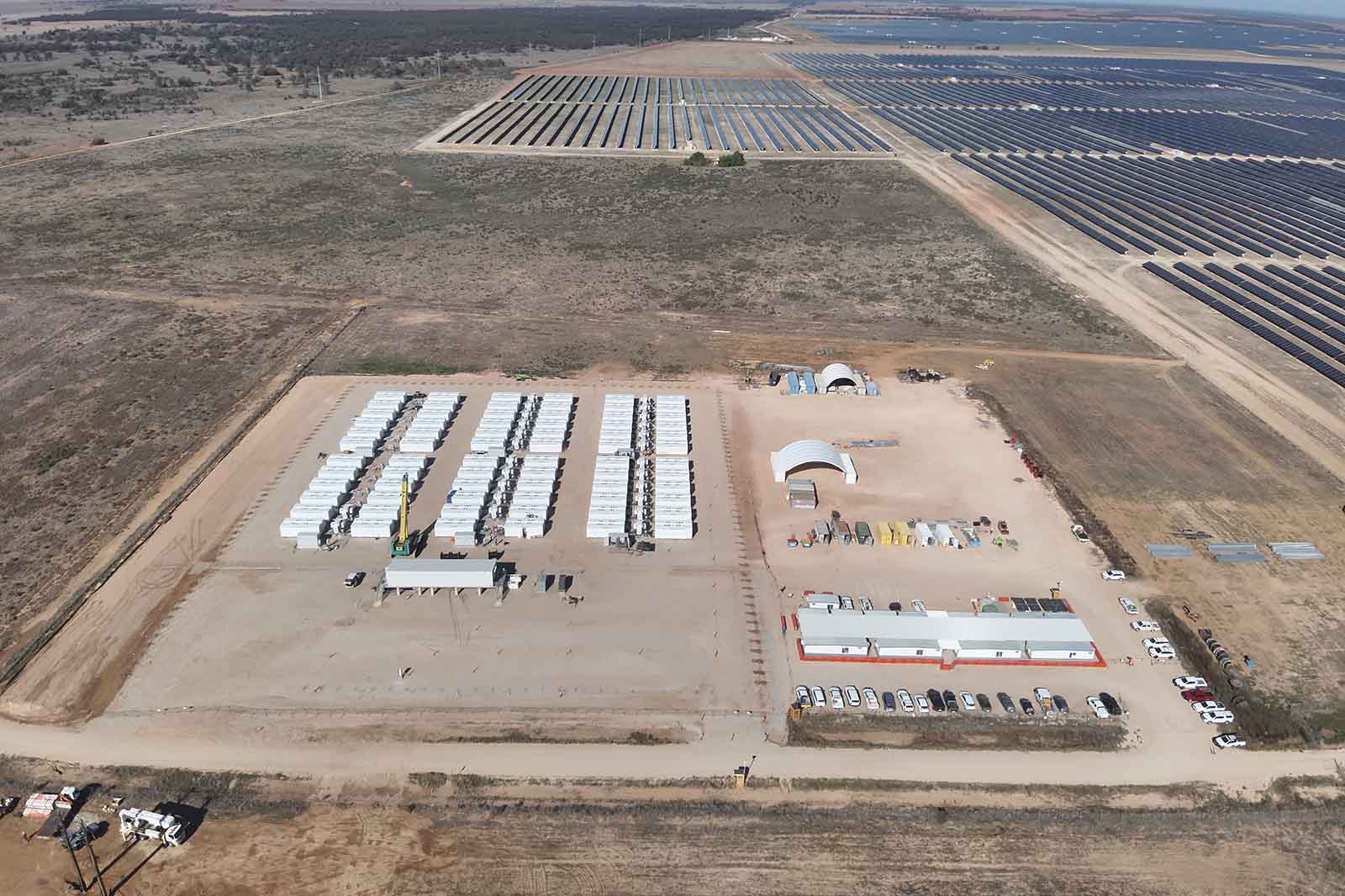 Aerial view of a dusty rectangular work compound with white modular units, parked trucks by beige structures, and distant dark solar panel arrays.