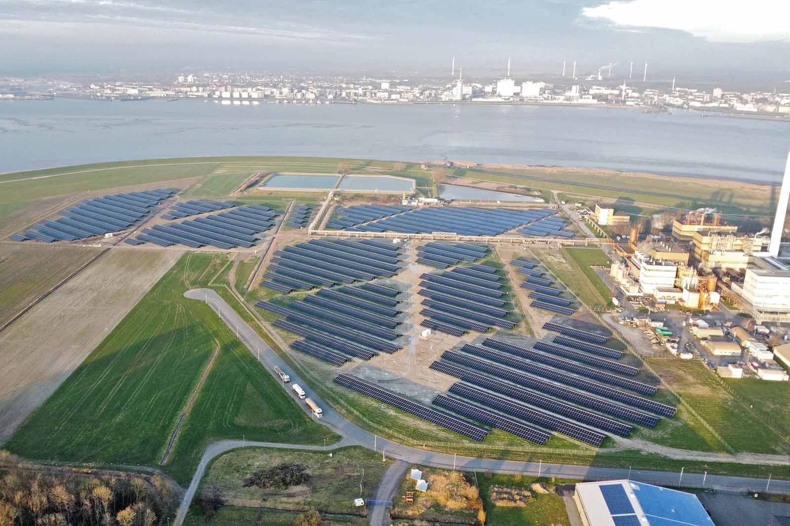 Aerial view of extensive blue solar panel arrays on flat green coastal land beside beige industrial buildings and a calm harbor across the water.
