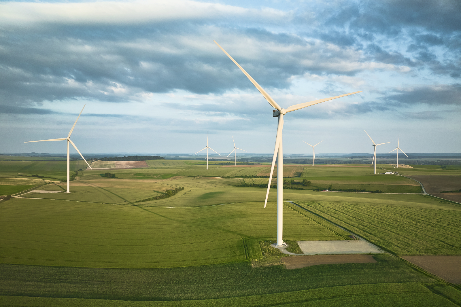 A tall white wind turbine stands in the foreground of patchwork green and brown agricultural fields under a blue-gray cloudy sky.