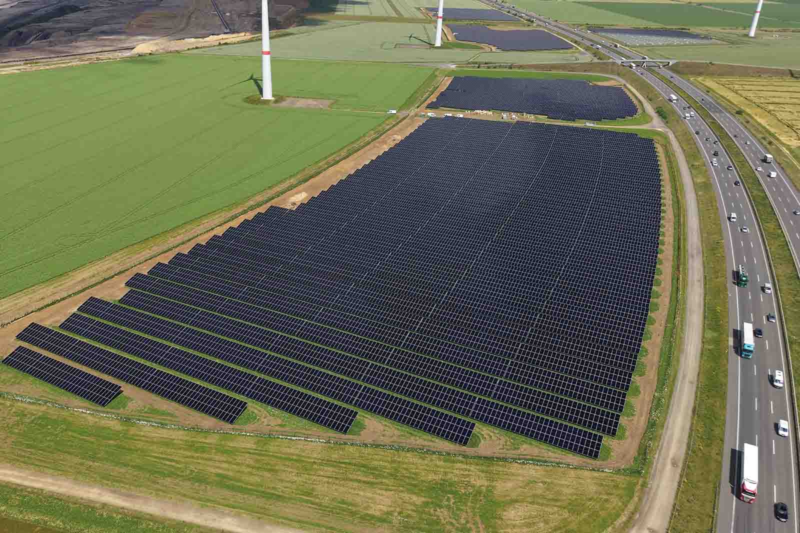 Großes Feld mit gleichmäßig angeordneten dunklen Solarmodulen im Vordergrund, umgeben von grünen Feldern, Windrädern und einer befahrbaren Autobahn rechts.
