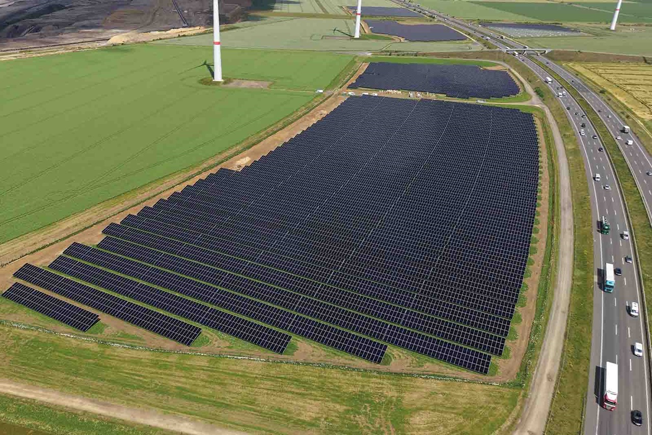 Großes Feld mit gleichmäßig angeordneten dunklen Solarmodulen im Vordergrund, umgeben von grünen Feldern, Windrädern und einer befahrbaren Autobahn rechts.