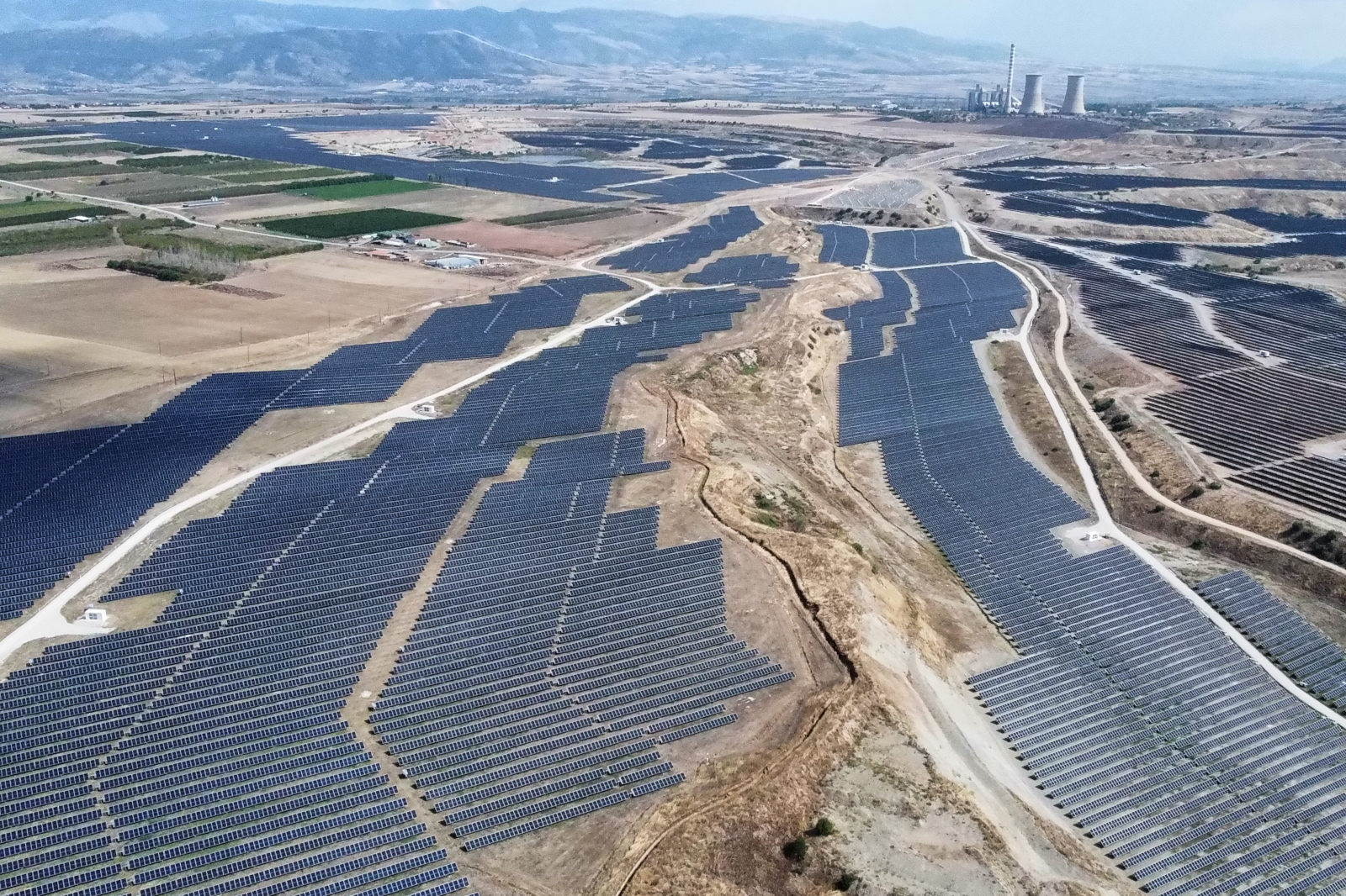 A vast solar farm fills the foreground with rows of dark blue panels across dry brown earth, intersected by dirt roads and distant cooling towers.