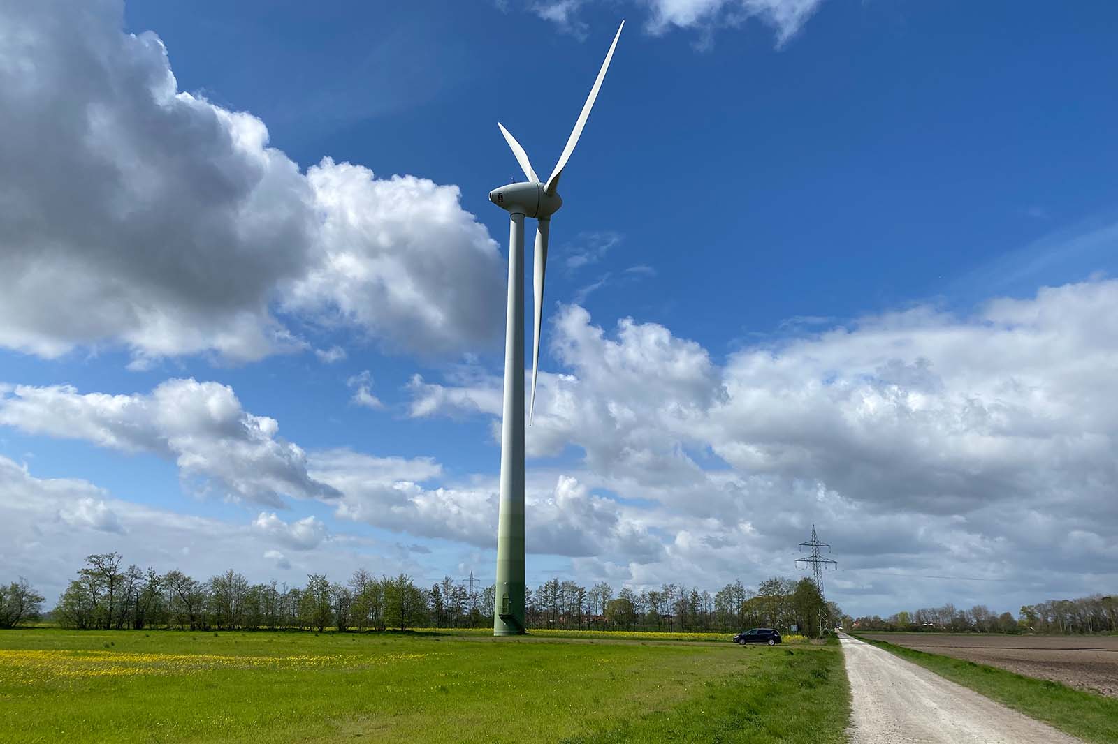 A tall white wind turbine rises from a green field under a blue sky with white clouds, a small dark car by a gravel road.