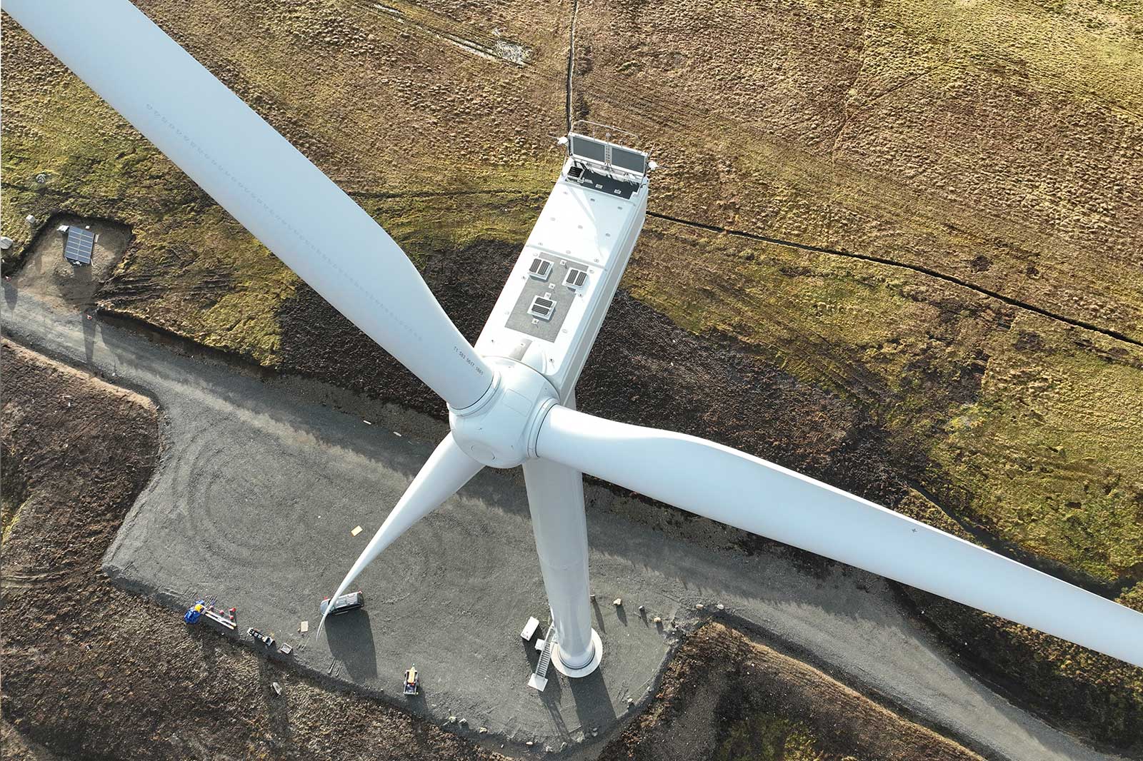 A white wind turbine viewed from above shows three long blades and a cylindrical tower on a gray gravel pad within brown grassy moorland.
