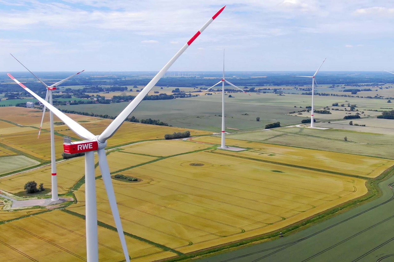 Ein großes weißes Windrad mit drei Rotorblättern und roter Gondel mit der Aufschrift „RWE“ über goldenen Getreidefeldern unter hellem blauen Himmel.