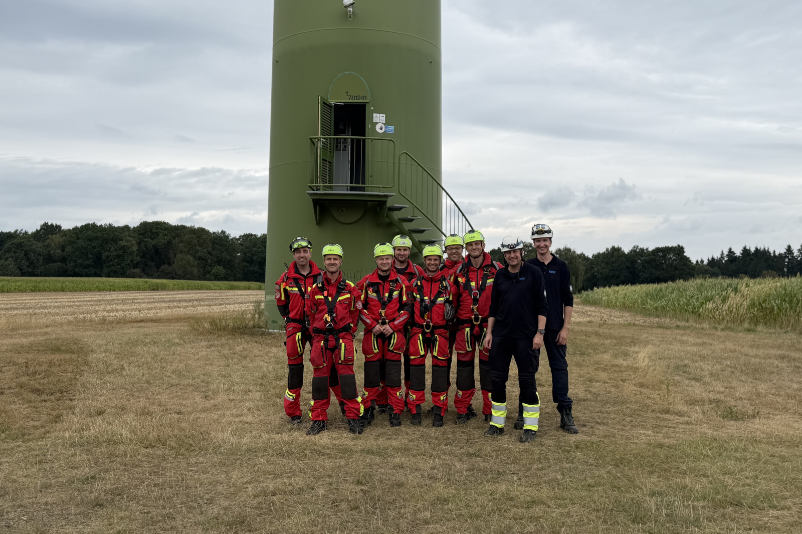 Neun Personen in roten Schutzanzügen und gelbgrünen Helmen stehen vor einem grünen metallischen zylindrischen Turm mit Außentreppe auf trockenem Feld unter grauem Himmel.