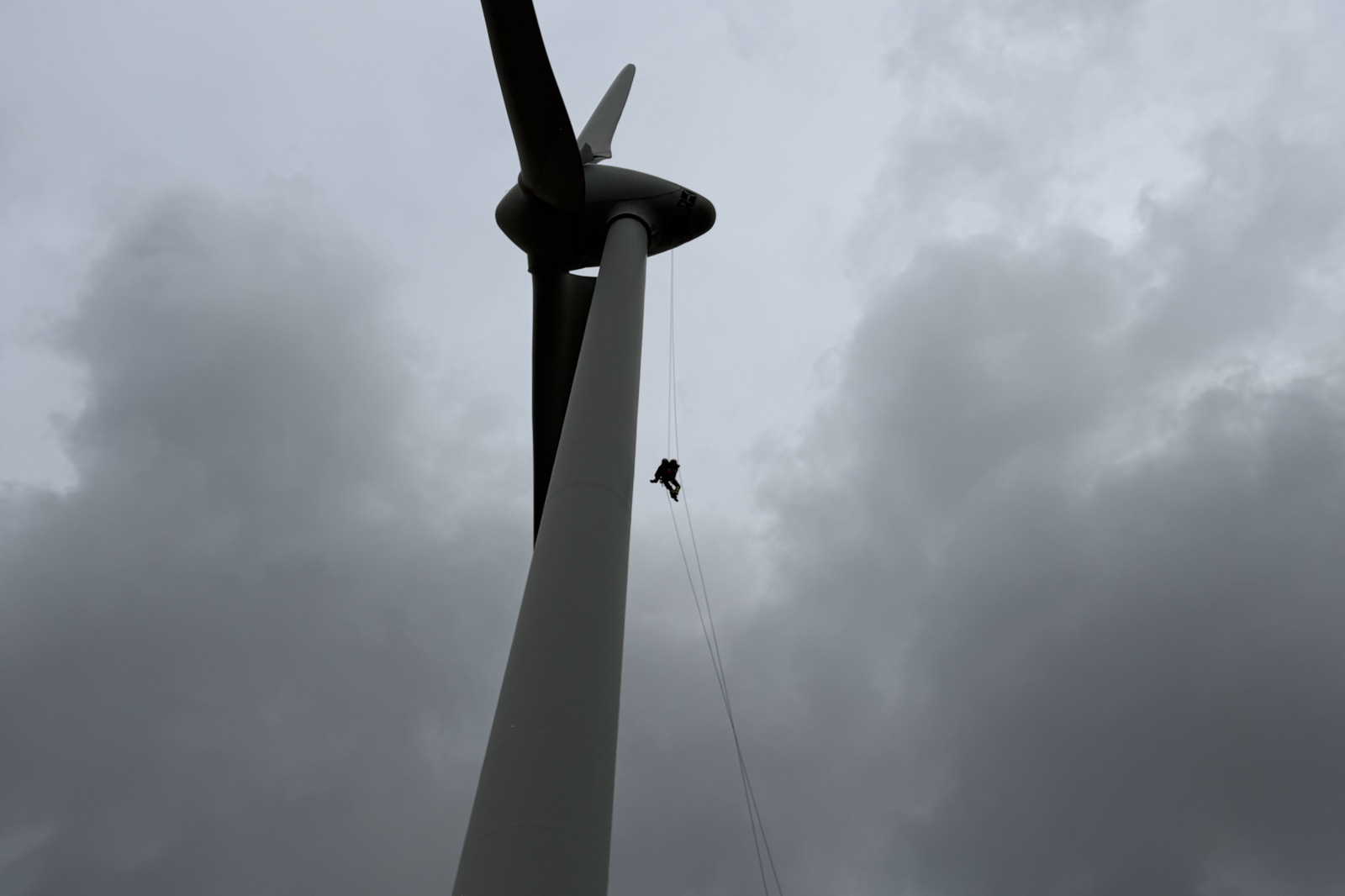 Ein weißer Windkraftturm mit Gondel und Rotorblatt vor einem düsteren, bewölkten Himmel, an dem eine Person an Seilen herabhängt.