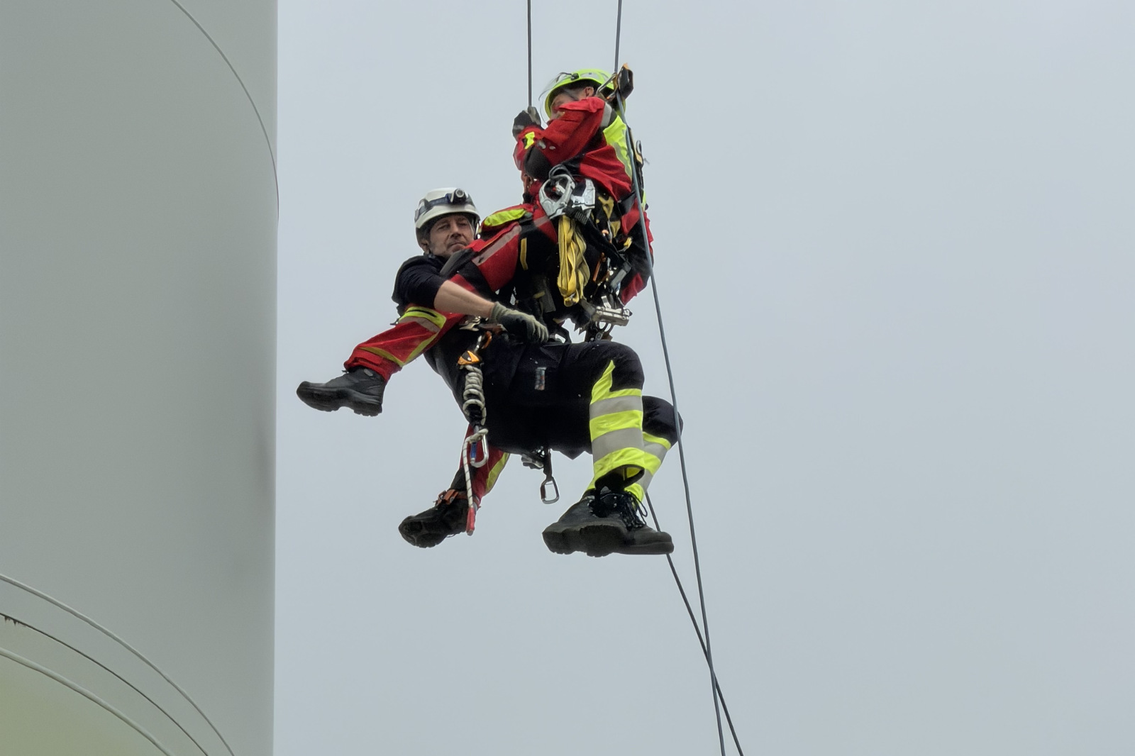 Zwei Rettungskräfte in roten und gelben Schutzanzügen und Helmen hängen in Sicherheitsgurten an Stahlseilen neben der weißen Turmwand vor grauem Himmel.
