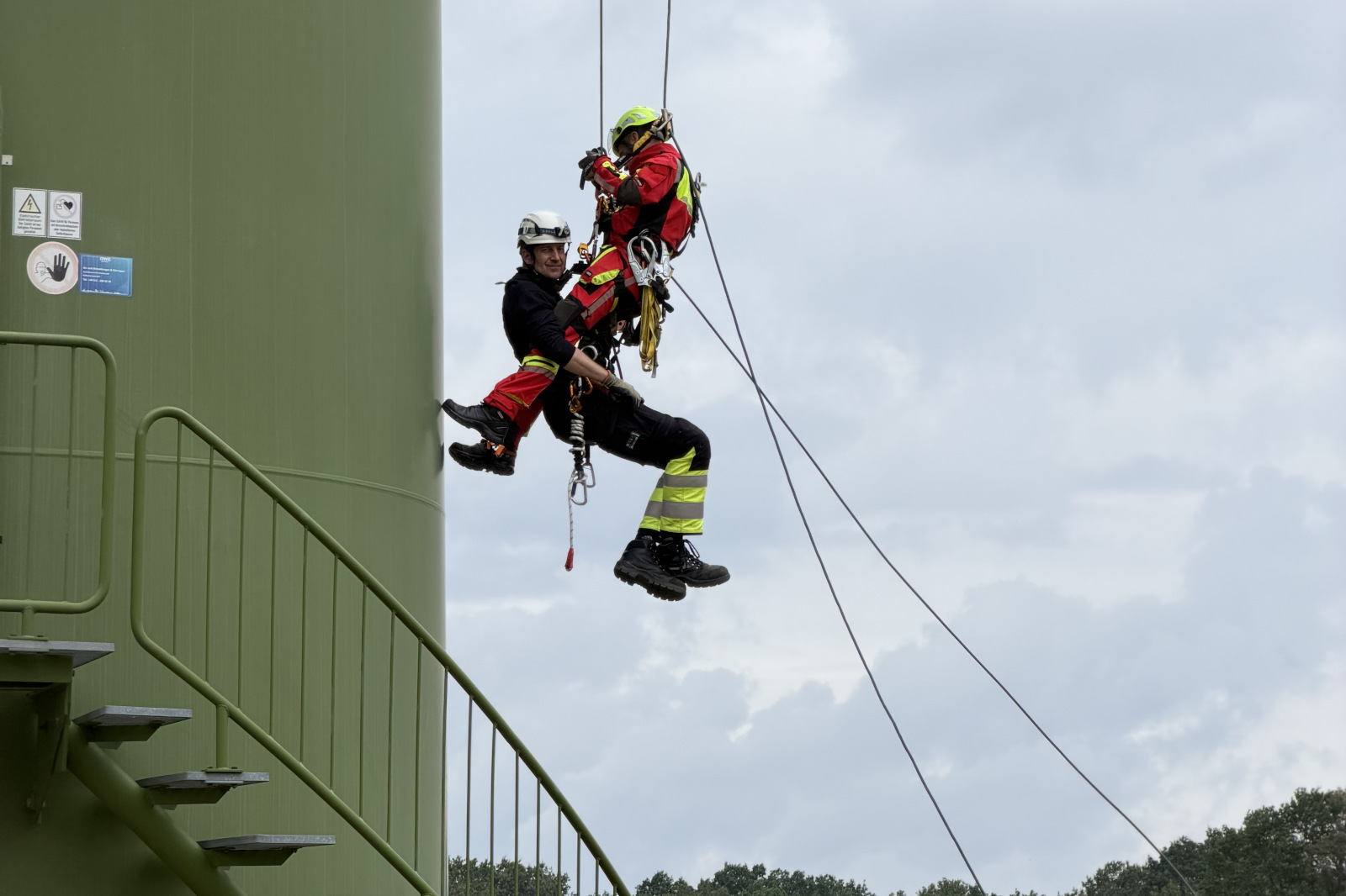 Zwei an Seilen hängende Einsatzkräfte, eine in roter Schutzkleidung, die andere in dunkler Uniform mit gelben Streifen, neben einem grünen Metallturm vor bedecktem Himmel.
