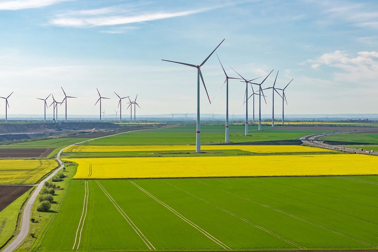 Hohe weiße Windräder ragen über ein Mosaik aus grünen und leuchtend gelben Feldern unter einem blassblauen Himmel und erzeugen eine ruhige ländliche Stimmung.