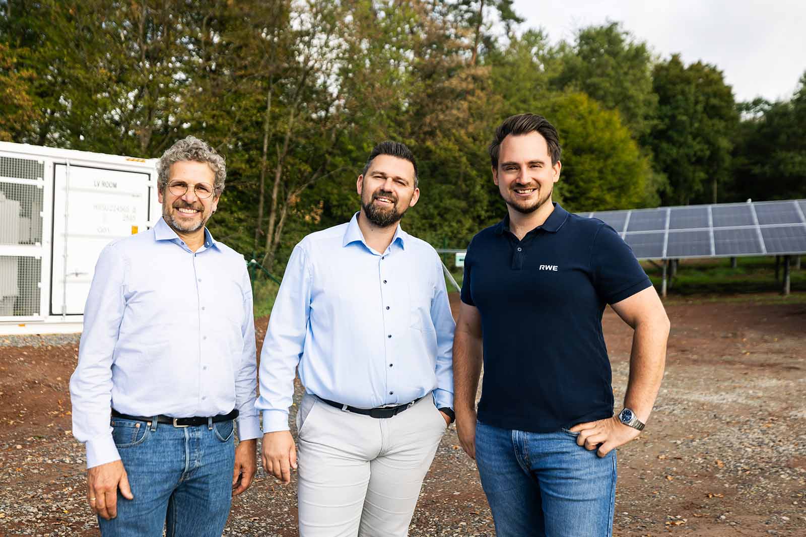 Drei lächelnde Männer stehen nebeneinander auf einem kiesigen Untergrund vor einem weißen Technikcontainer und einer Solaranlage, zwei in hellblauen Hemden, einer in dunkelblauem Polo.