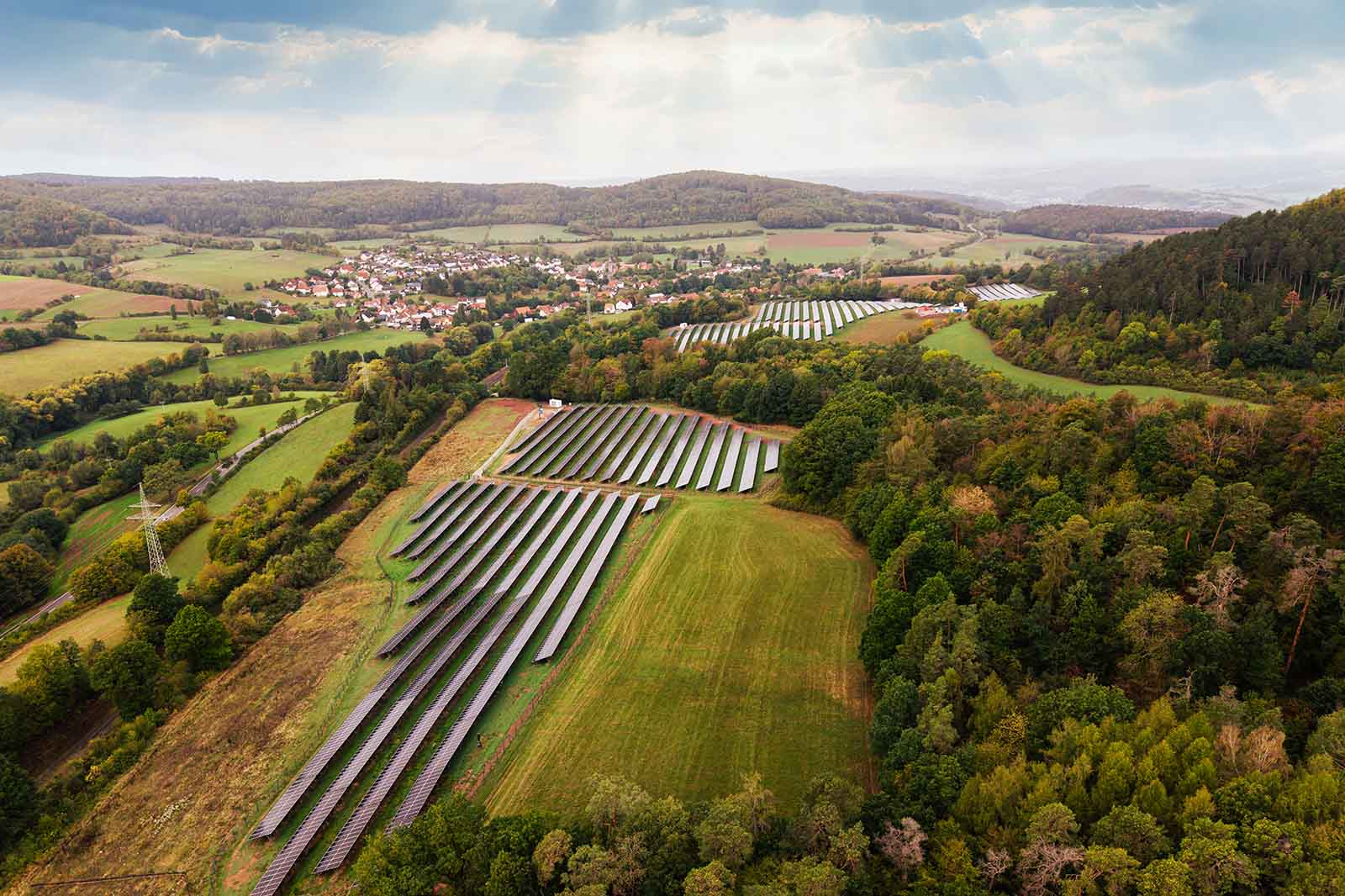 Eine Reihe glänzender Solarmodule steht auf grünem Feld im Vordergrund, umgeben von herbstlich gefärbten Wäldern, Hügeln und einem Dorf unter einem hellen Himmel.
