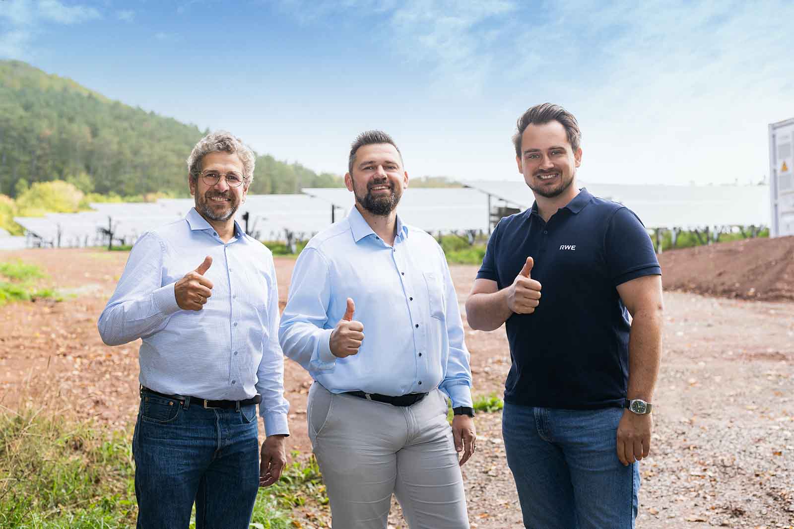 Drei lächelnde Männer stehen vor einer Reihe glänzender Solarmodule auf einem Schotterweg, zwei tragen hellblaue Hemden, einer ein dunkles Poloshirt, alle zeigen Daumen hoch.