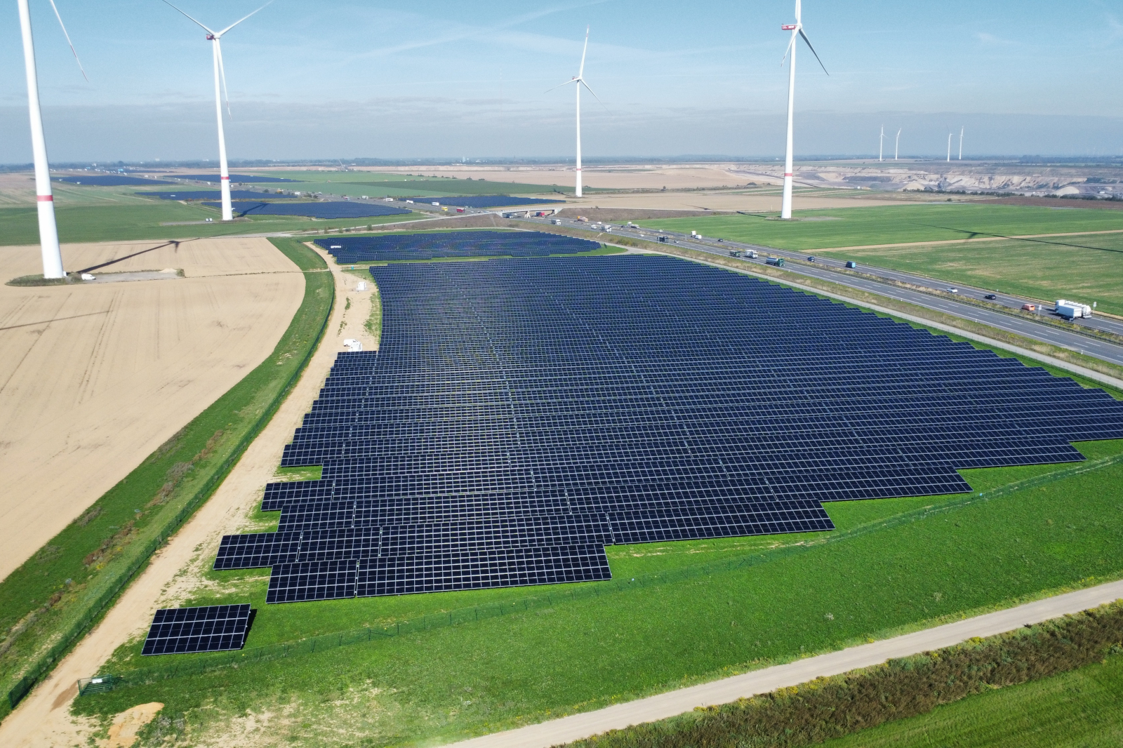 Großes Feld mit dunklen, glänzenden Solarmodulen im Vordergrund, flankiert von grünen Wiesen und beigen Ackerflächen, dahinter weiße Windkraftanlagen unter hellem, blassem Himmel.
