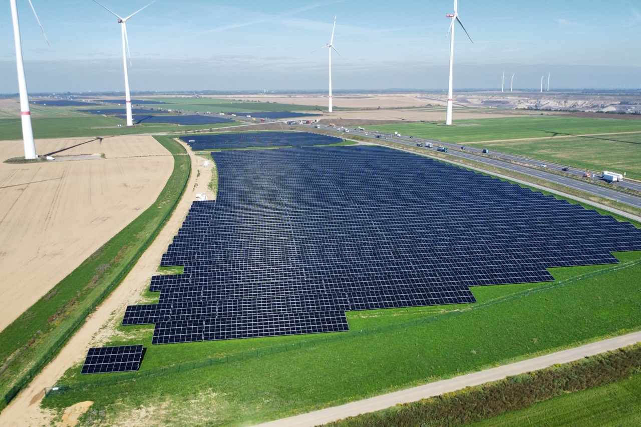 Großes Feld mit dunklen, glänzenden Solarmodulen im Vordergrund, flankiert von grünen Wiesen und beigen Ackerflächen, dahinter weiße Windkraftanlagen unter hellem, blassem Himmel.