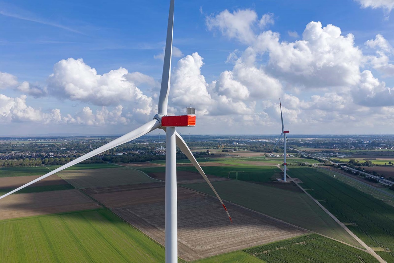Eine weiße Windkraftanlage mit roter Gondel und langen Rotorblättern dominiert den Vordergrund, dahinter weite grüne und braune Felder unter blauem Himmel mit weißen Wolken.